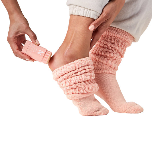 Person applying Overnight Heel Repair Set balm to heel while wearing soft pink knit socks on white background