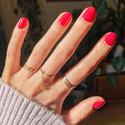 Hand with Pomodoro nail polish in vibrant pinky-tomato red on natural nails adorned with delicate gold rings