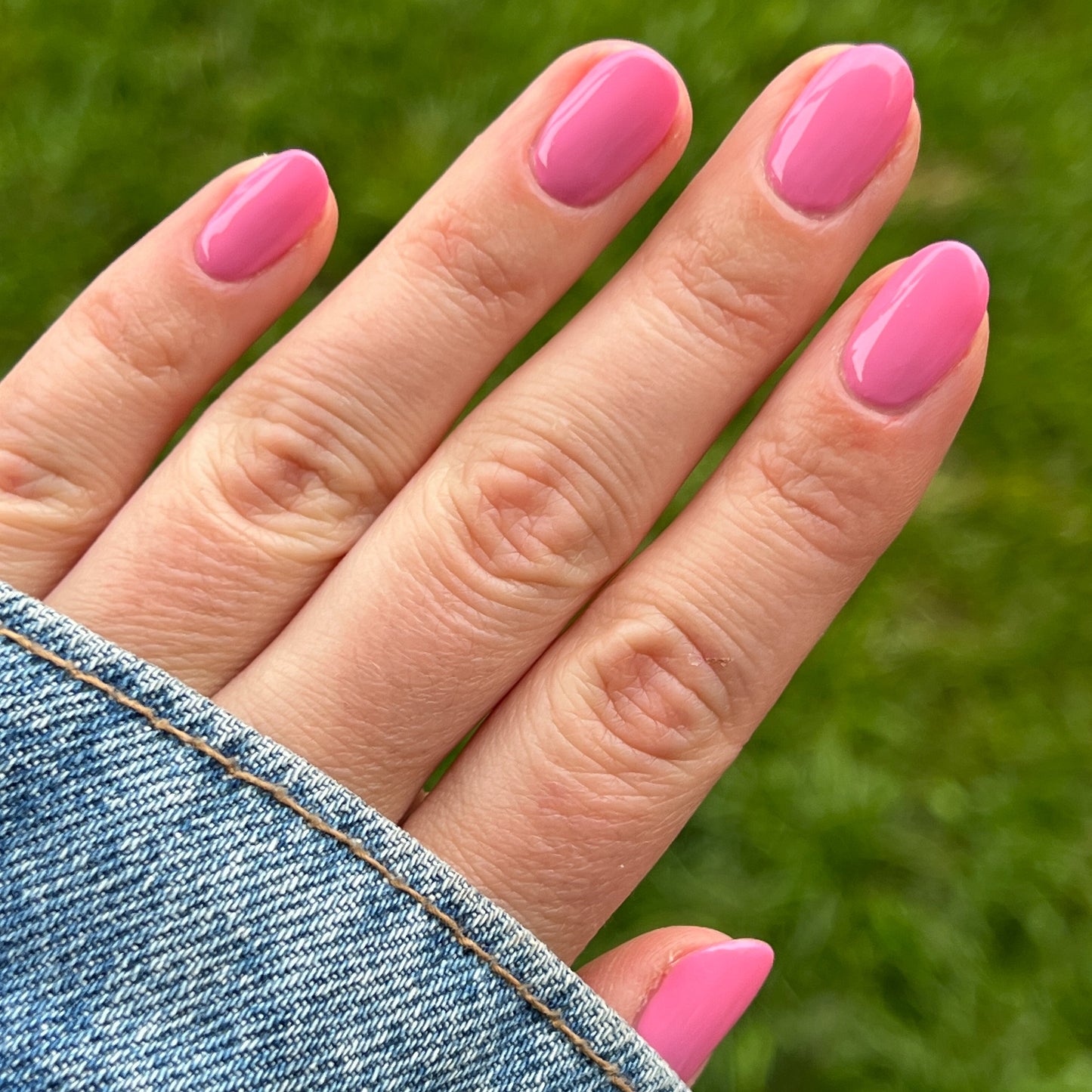 Hand with nails painted in bright pink Taffy gel polish shown outdoors with denim jacket sleeve and green grass background