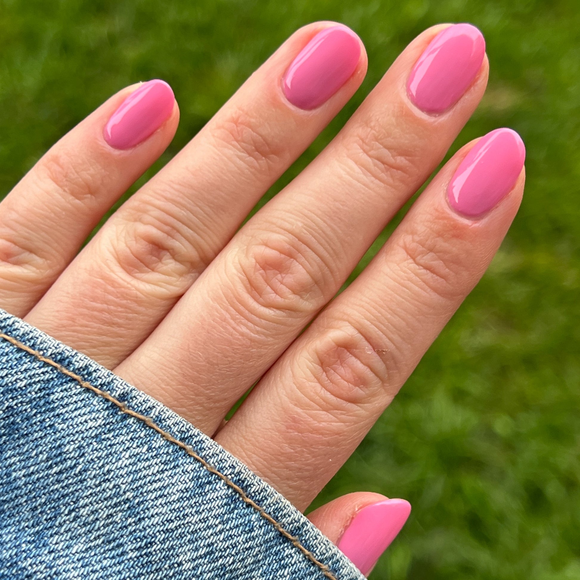 Hand with nails painted in bright pink Taffy gel polish shown outdoors with denim jacket sleeve and green grass background