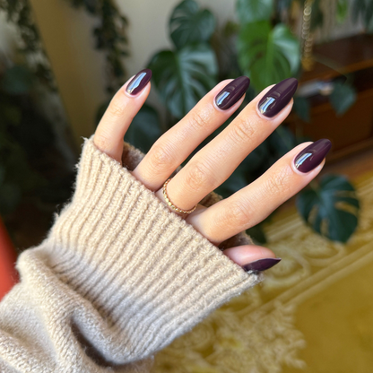 Hand with medium skin tone showing glossy nails in Covet - Gel, a cool dark plum hue, wearing a cozy beige sweater sleeve