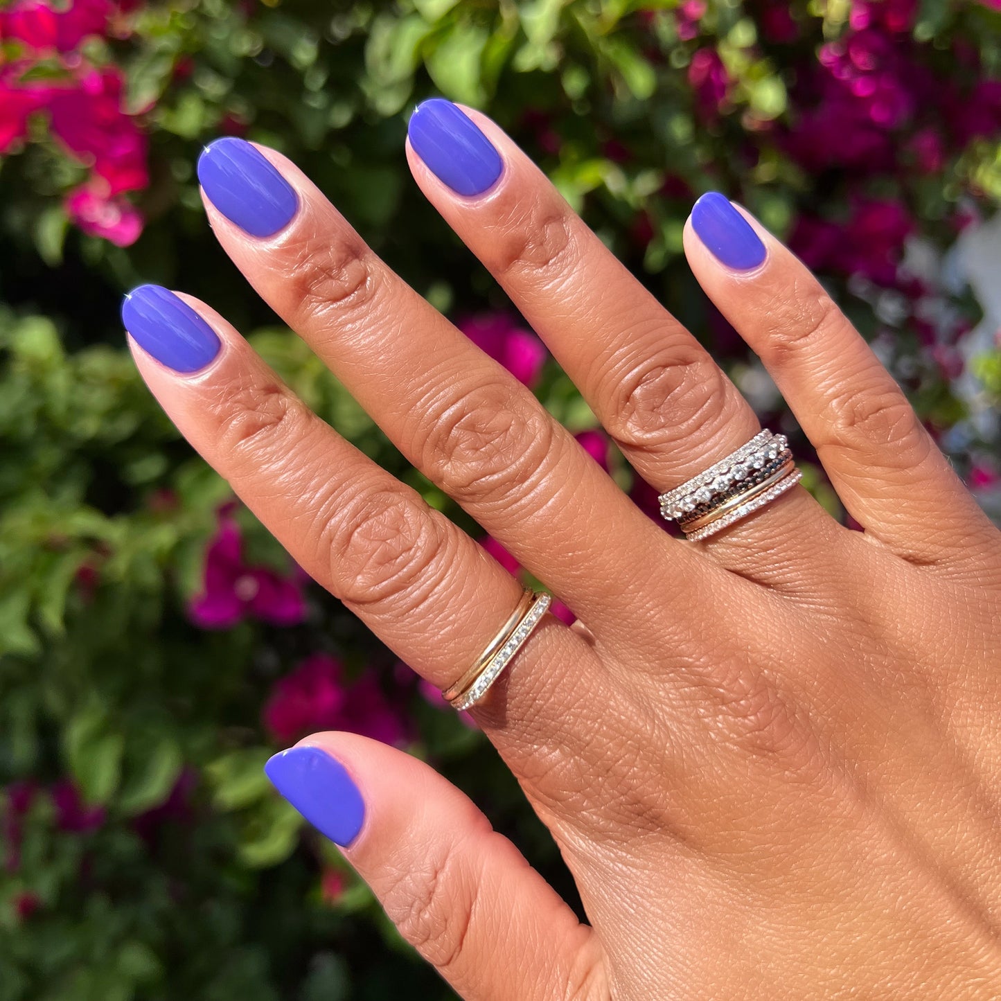 Hand with nails polished in vibrant deep purple House of O&J polish wearing layered rings outdoors with pink flowers background