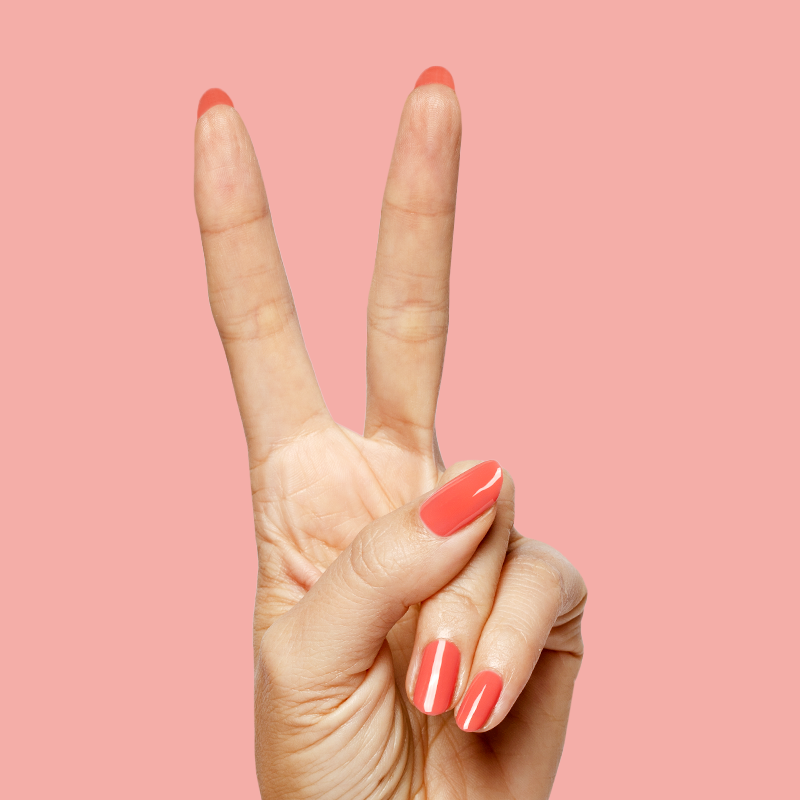 Hand showing peace sign with nails polished in glossy coral shade using Field Day - Gel on soft pink background