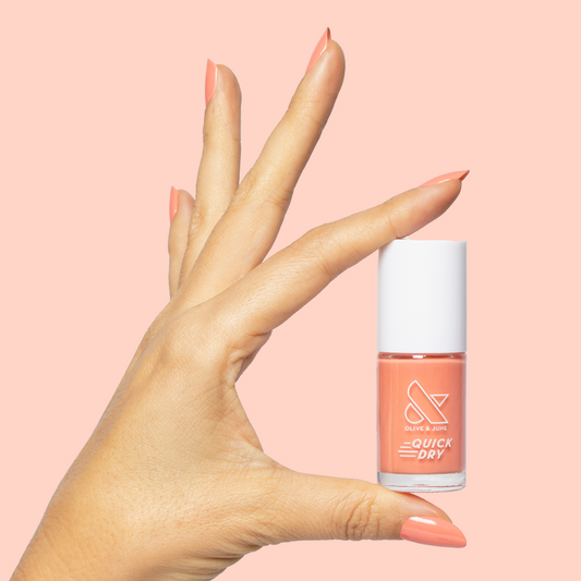 Hand holding Frolic bright orangey coral quick dry gel polish bottle with matching mani on soft peach background