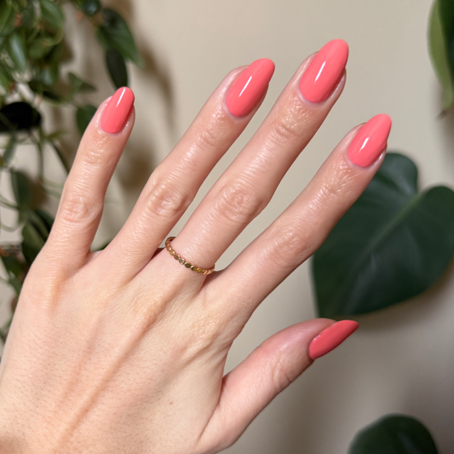 Hand with glossy almond-shaped nails painted in bright orangey coral Frolic gel polish against blurred green plants background