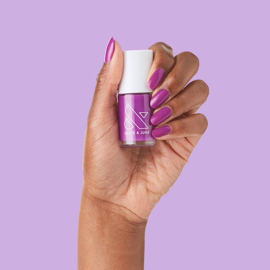 Hand holding Grape Soda nail polish bottle with matching bright purple mani and smooth glossy finish on neutral background