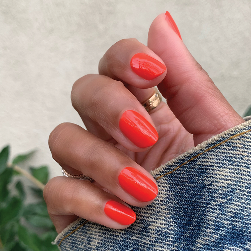 Hand with Lava polish in bright spicy orange red showing a smooth glossy manicure against denim fabric background