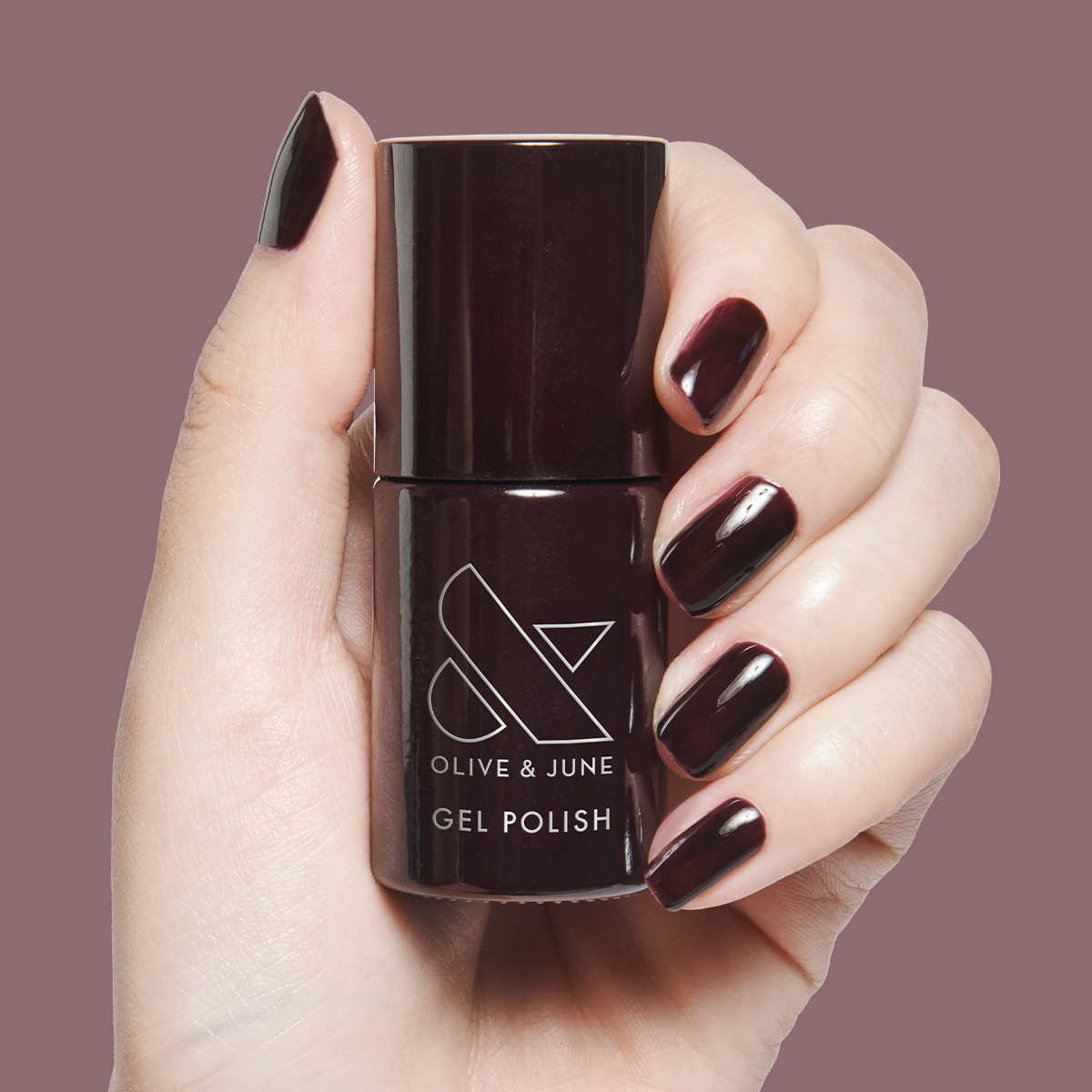 Hand holding bottle of Covet - Gel gel polish with nails painted in a deep dark plum shade on muted mauve background