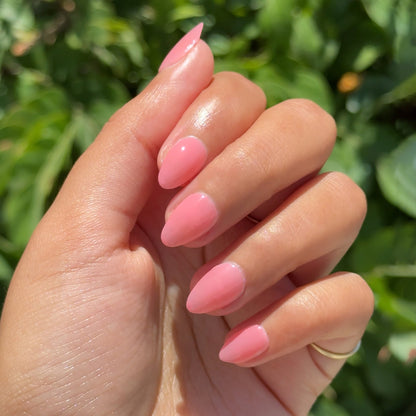 Hand with medium almond-shaped nails polished in glossy sheer coral pink using Papaya Gloss gel polish outdoors with greenery background