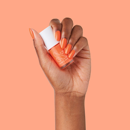 Hand with nails painted in bold tangerine You're a 10 gel polish holding matching polish bottle on orange background