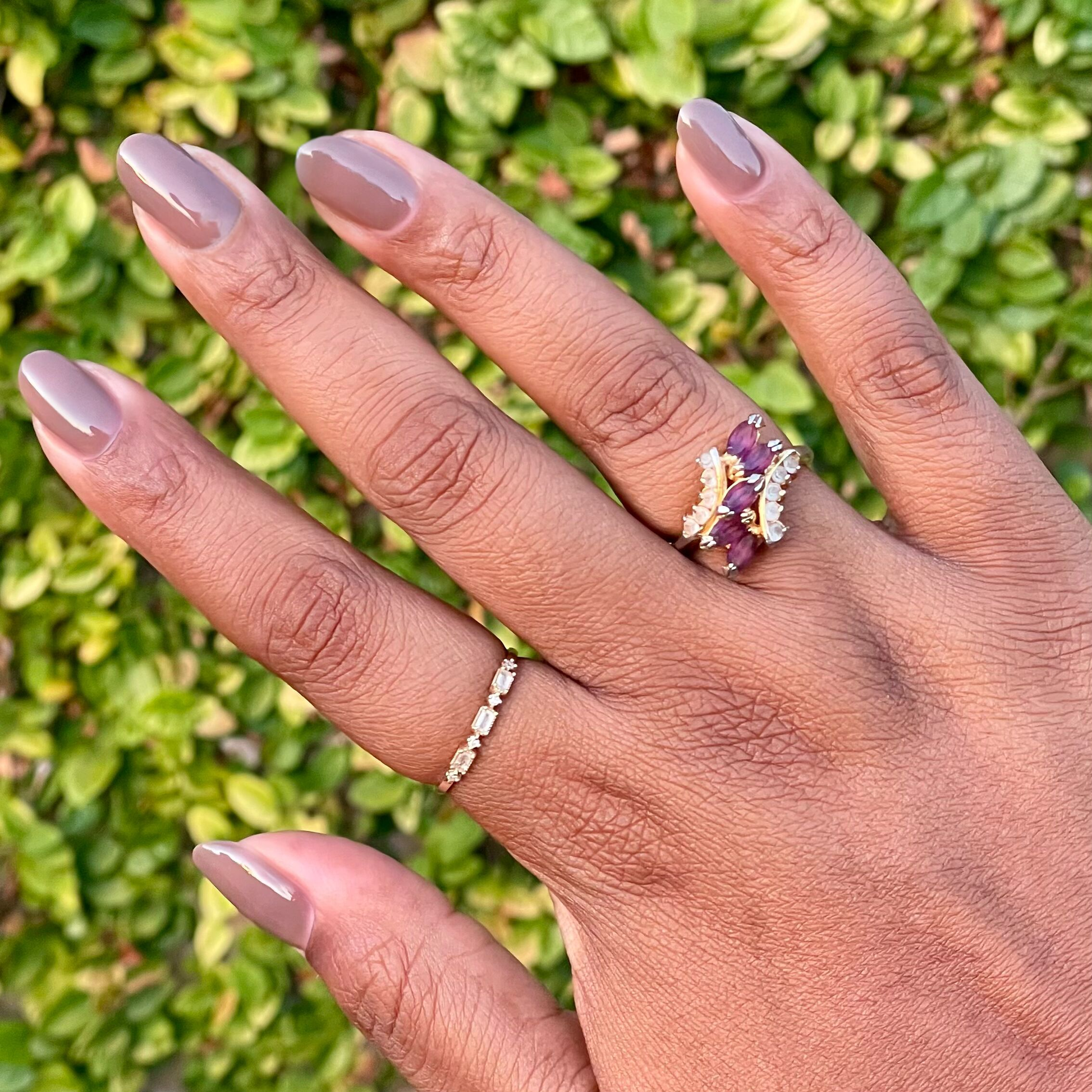 Hand with nails painted in Fawn - Gel, a muted brown neutral gel polish, adorned with delicate gold rings against a green background