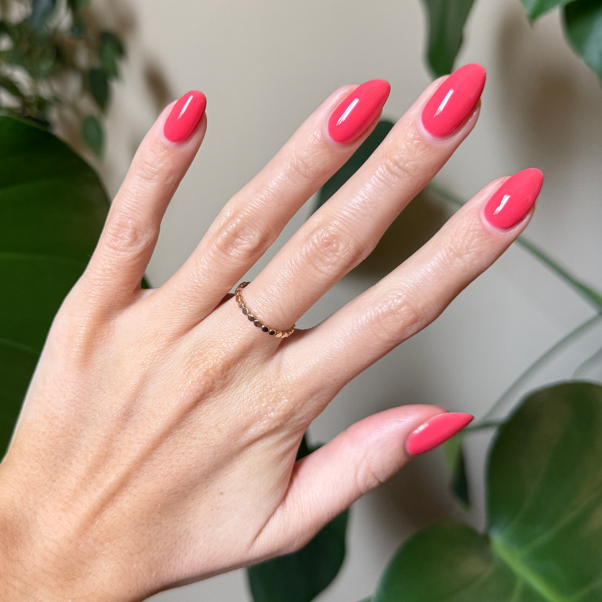 Hand with almond-shaped nails polished in juicy watermelon red using Field Day - Gel against natural leafy background