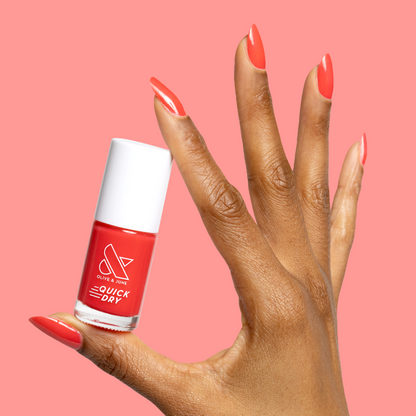 Hand with nails painted in glossy Fragola quick dry polish holding matching bottle against a coral background
