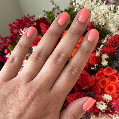 Hand with short rounded nails painted in bright orangey coral Frolic gel polish over dried colorful flowers background