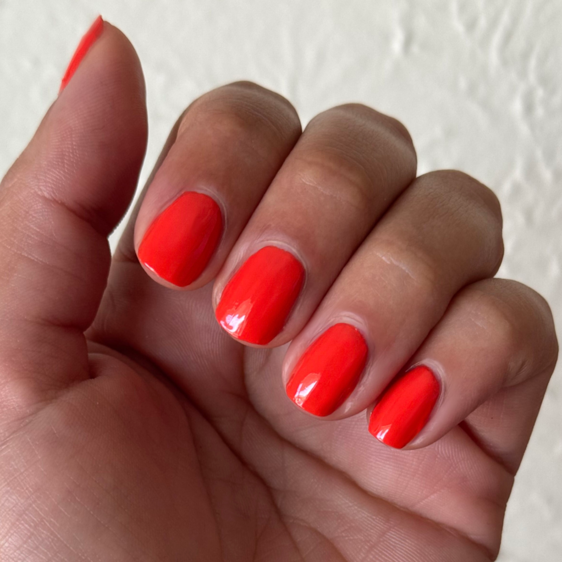 Hand with rounded nails painted in bright orange-red Snapdragon quick dry polish against neutral light background